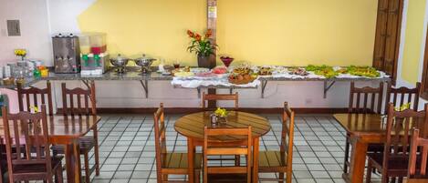 A dining area with a round wooden table and chairs, a tiled floor, a yellow wall, and a counter with food and drinks.