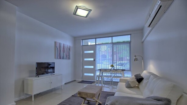 Een moderne woonkamer met een flatscreen-tv, een witte bank, een klein houten tafeltje en een eethoek met een witte tafel en stoelen.
