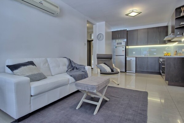A modern living room with a white sofa, a grey armchair, and a small table. A kitchen with dark cabinets and a microwave is visible in the background.