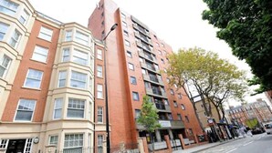 Ein mehrstöckiges Wohnhaus mit Hauseingang an der Ecke, einem Balkon im Erdgeschoss und einem Baum davor.