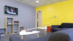 Ein Zimmer mit Fernseher an der Wand, einem Bücherregal, einem Schreibtisch, einer Couch und einer gelben Wand.