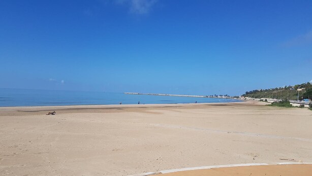Een breed zandstrand met een helderblauwe lucht, een rustige zee en een pier in de verte.