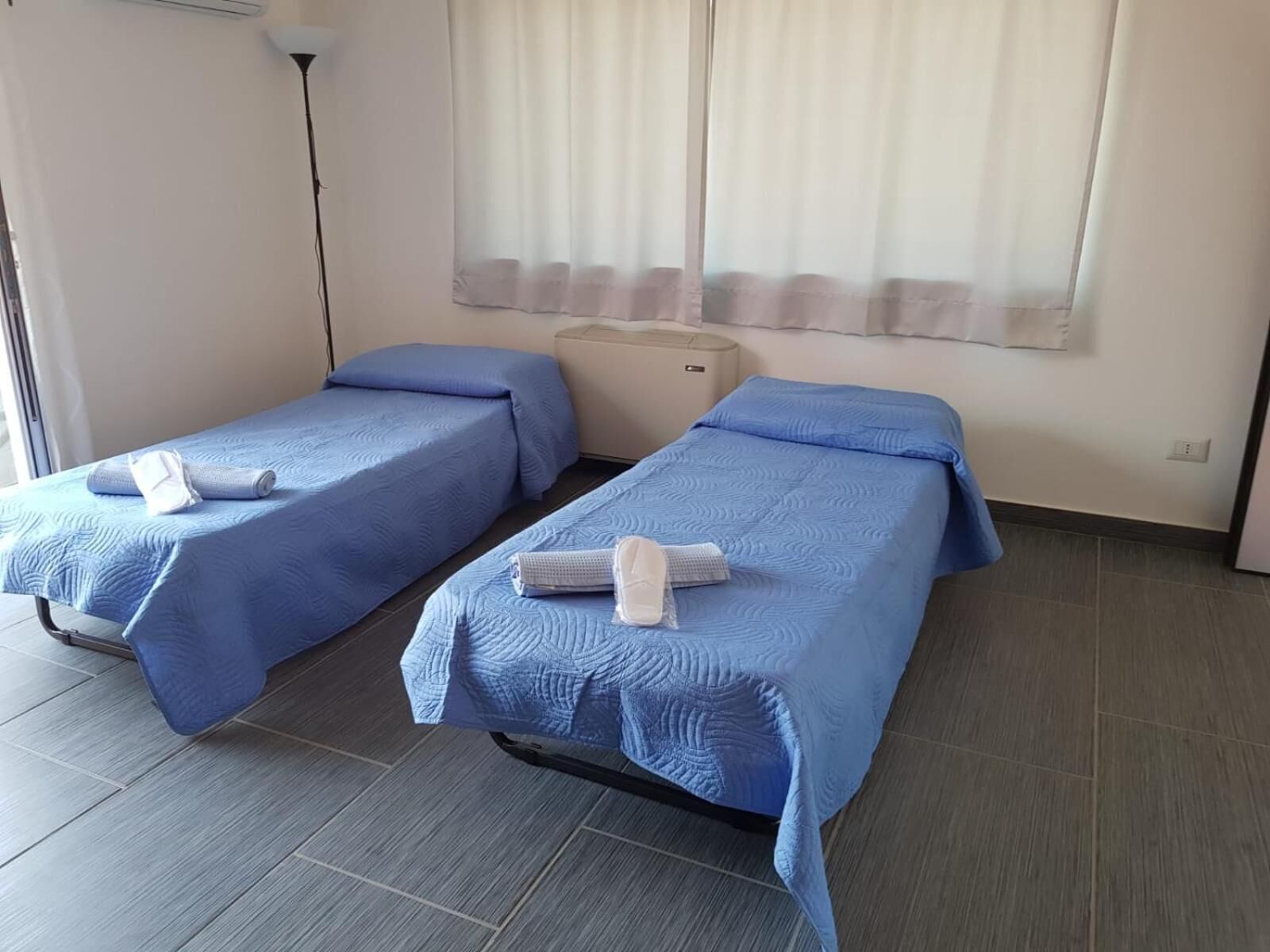 Dos camas individuales con ropa de cama y toallas azules, una mesita de noche, una lámpara y una ventana con cortinas.
