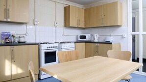 A compact kitchen with wooden cabinets, a microwave, a stove, and a dining table. - Wood Green Hall - Campus Accommodation (London)