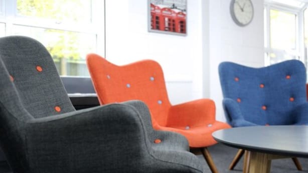 A seating area with three chairs in grey, orange, and blue, a round table, and a clock on the wall. - Wood Green Hall - Campus Accommodation (London)