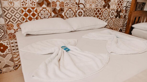 A neatly made bed with white linens and a blue tag, set against a patterned tile wall. - Pousada Armação das Baleias (Penha)