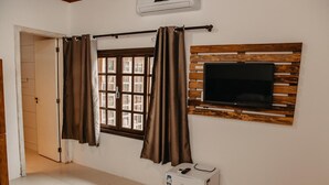 A room with a window, curtains, a television mounted on a wooden panel, and a small refrigerator. - Pousada Armação das Baleias (Penha)