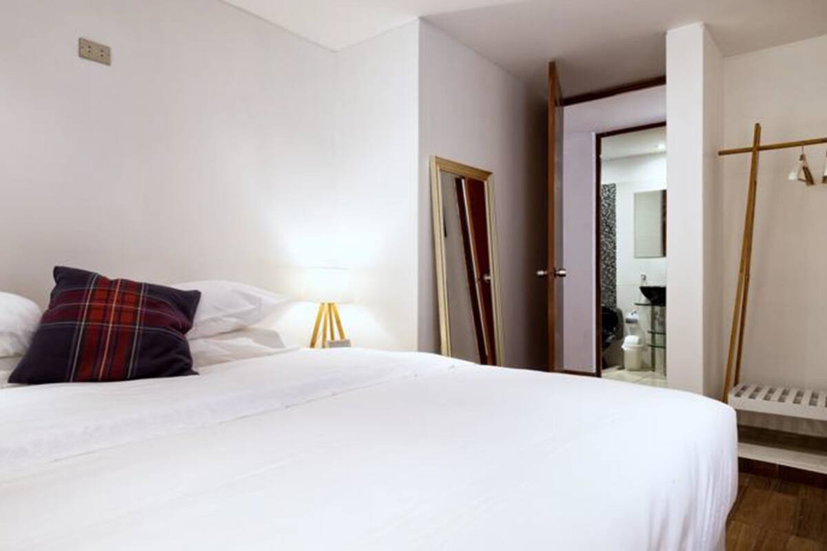 A hotel room with a large bed, a bedside table with a lamp, a wardrobe, and a bathroom visible through an open door.