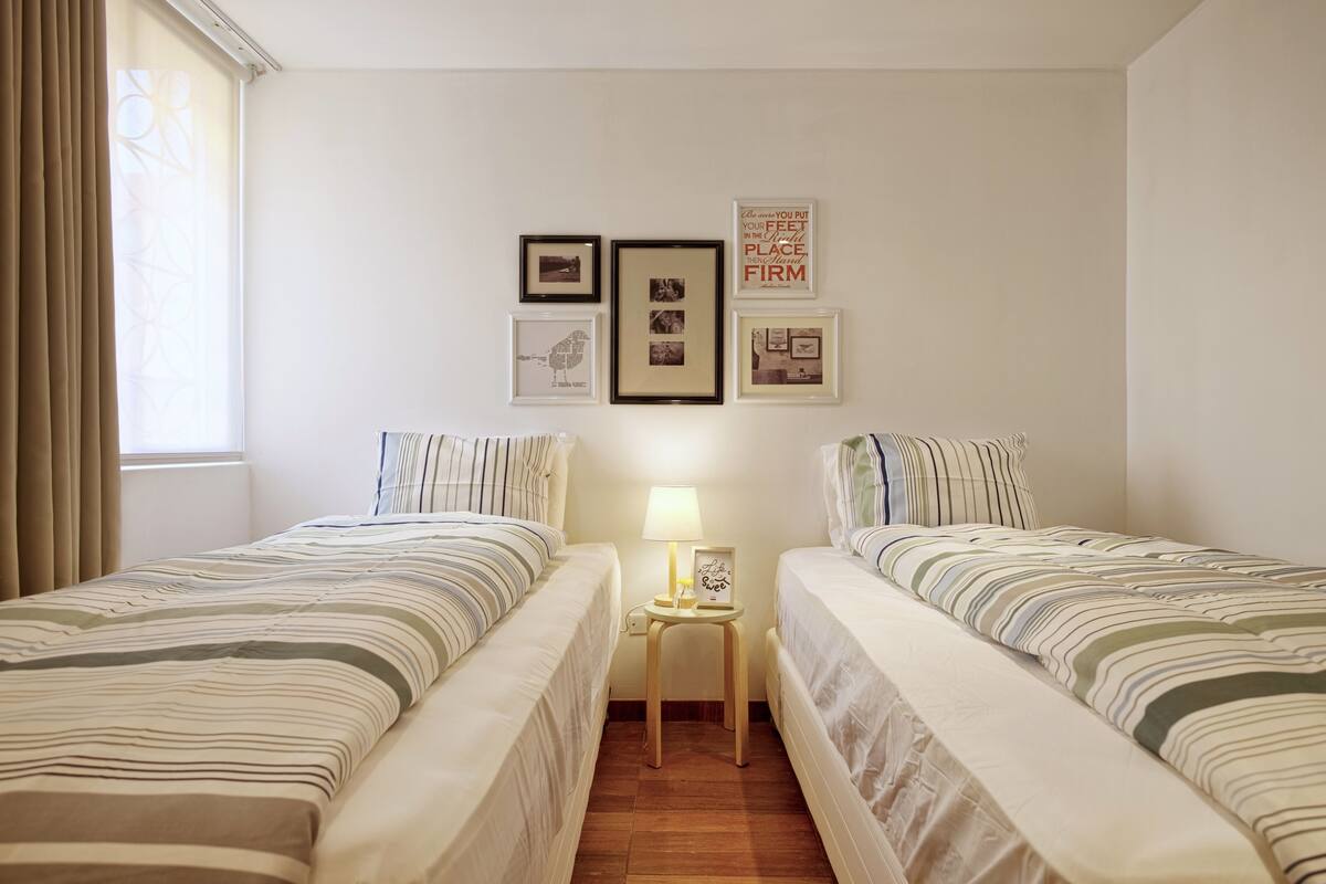 A hotel room with two beds, a small table with a lamp, and framed pictures on the wall.