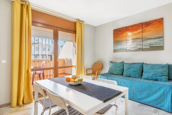 A room with a dining table, a sofa, a painting of a sunset, and a view through a window.