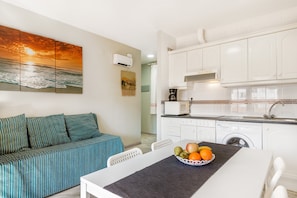 A modern kitchen with white cabinets, a dining table with a fruit bowl, a sofa with striped cushions, and a wall-mounted air conditioner.