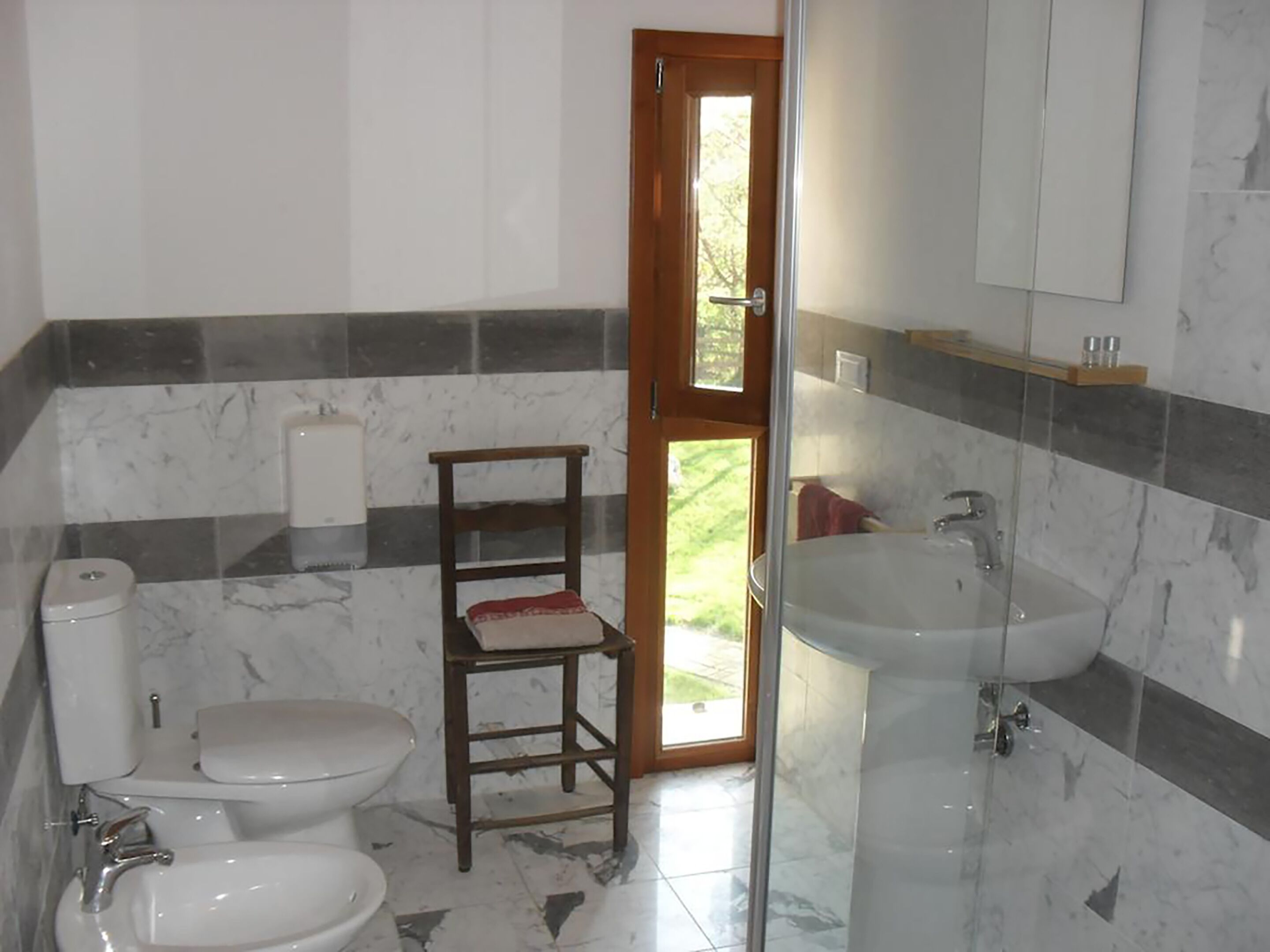 Baño con inodoro, bidé y lavamanos, baldosas de mármol y una silla de madera.