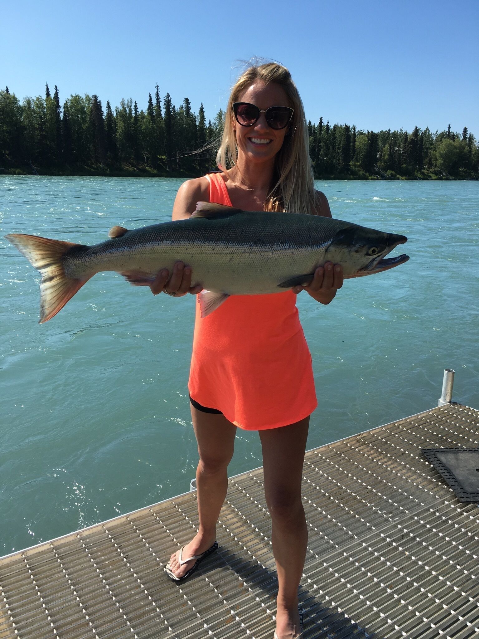 A person holding a fish on a dock with a lake and forest in the background.