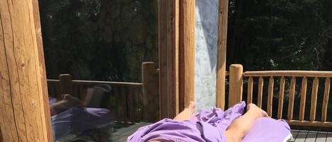 A wooden deck with a massage table and a chair, surrounded by wooden railings and a glass enclosure.
