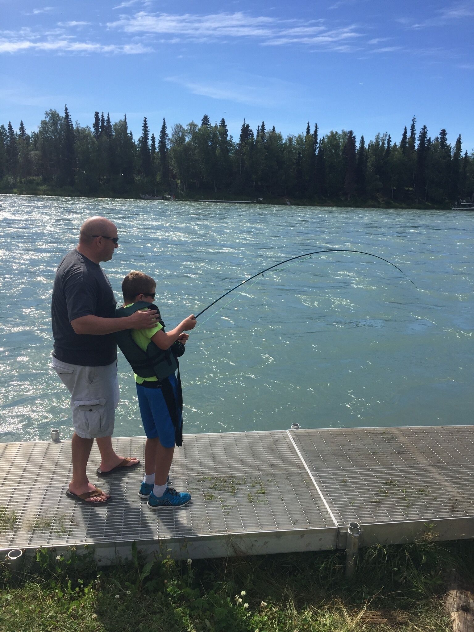 A man and a child fishing by a calm lake with a forest in the background.