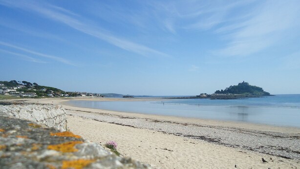 A beach with a rocky foreground, a sandy shore, and a distant hill with a structure on top. - Mounts Bay Caravan Park (Marazion)