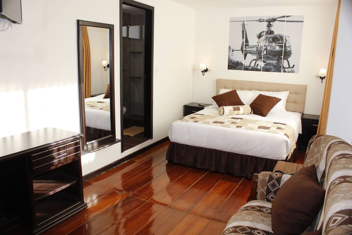 A hotel room with a bed, a nightstand, a mirror, a sofa, and a wooden floor.
