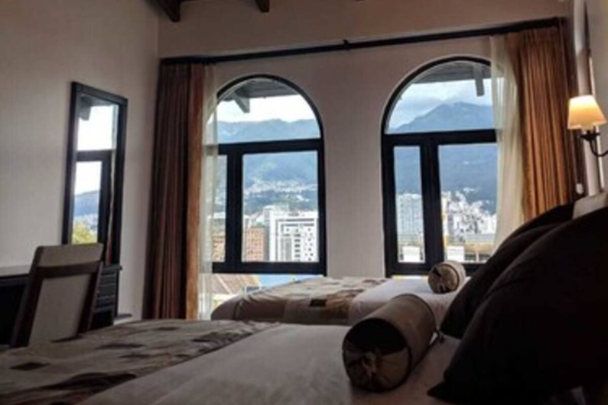 A hotel room with a large bed, two windows with arched tops, and a view of a cityscape and mountains.
