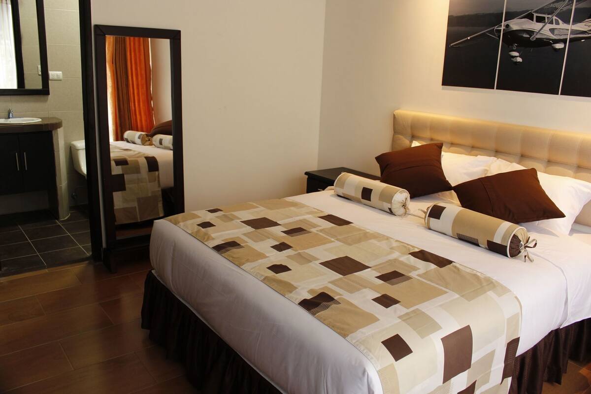 A neatly made bed with a patterned bedspread, a bedside table, a mirror, and a wall-mounted picture.