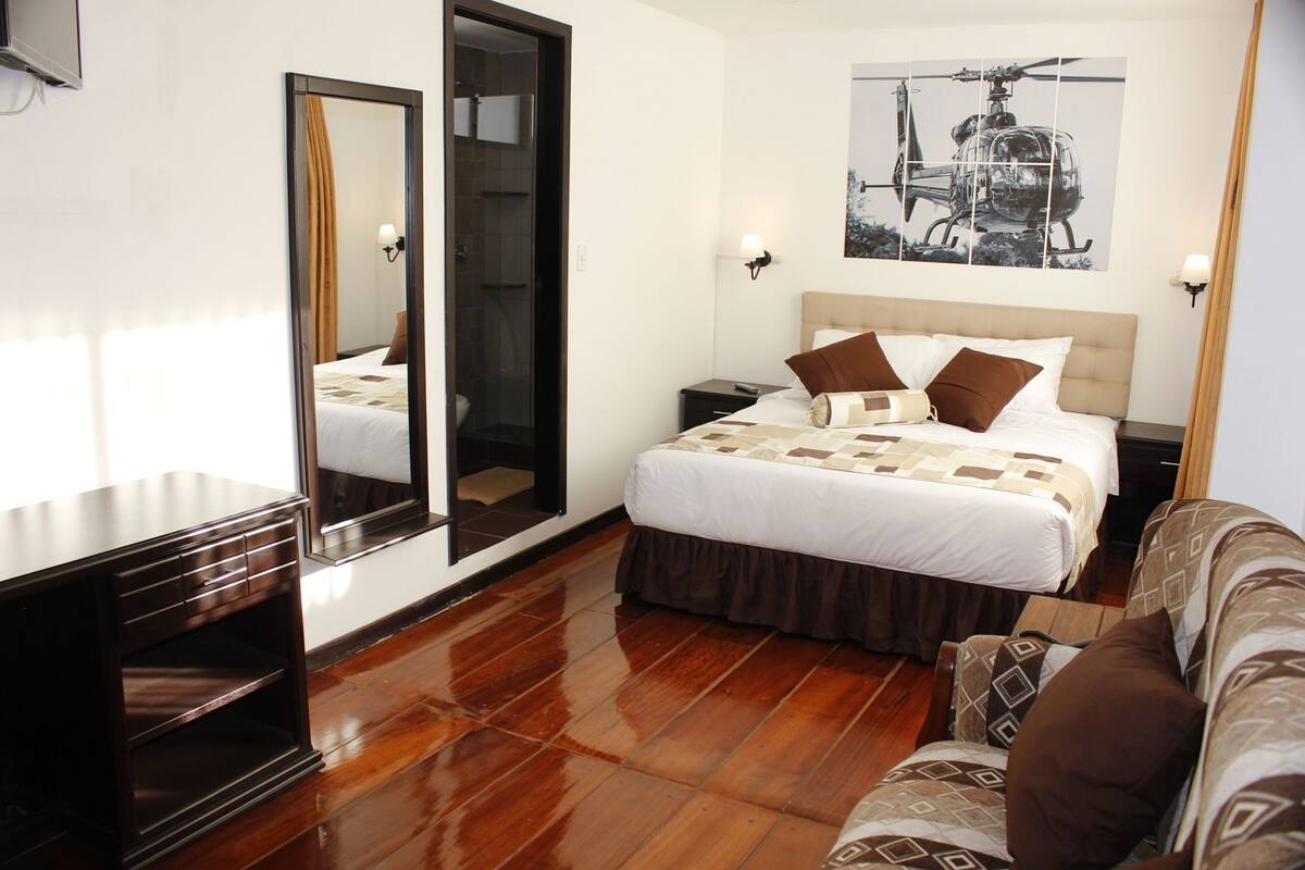 A hotel room with a bed, a sofa, a TV, a mirror, and a painting on the wall.