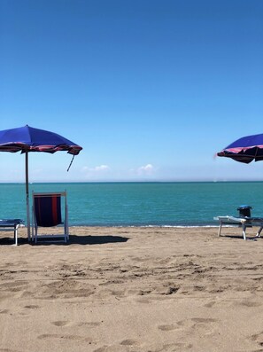 Ein Strand mit zwei Sonnenschirmen und Liegestühlen, bewölkter Himmel und ruhigem Meer.