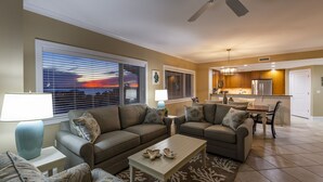 A living room with a sofa, coffee table, and a view of the sunset through the window. - Meridian 601 West by Seaside Vacations & Sales (Ocean City)