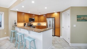A modern kitchen with wooden cabinets, a stainless steel refrigerator, a microwave, and a granite countertop. There are three light blue barstools and a small island with a sink. - Meridian 601 West by Seaside Vacations & Sales (Ocean City)