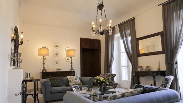 A living room with a sofa, armchairs, a coffee table, and a chandelier. There are lamps on the wall, a mirror, and a door in the background. - Antica Dimora La Portazza (Marsala)