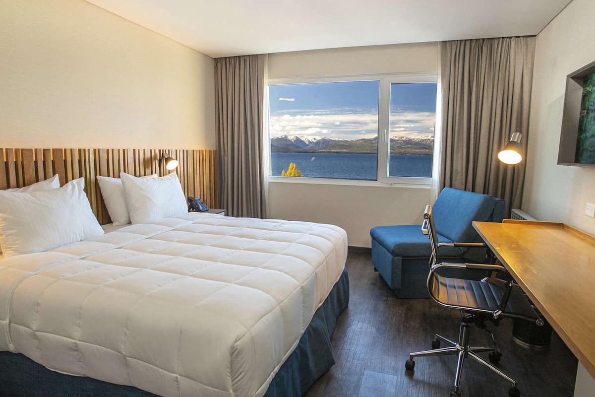 Habitación de hotel con una cama grande, un escritorio, una silla y vistas a las montañas y al agua a través de la ventana.