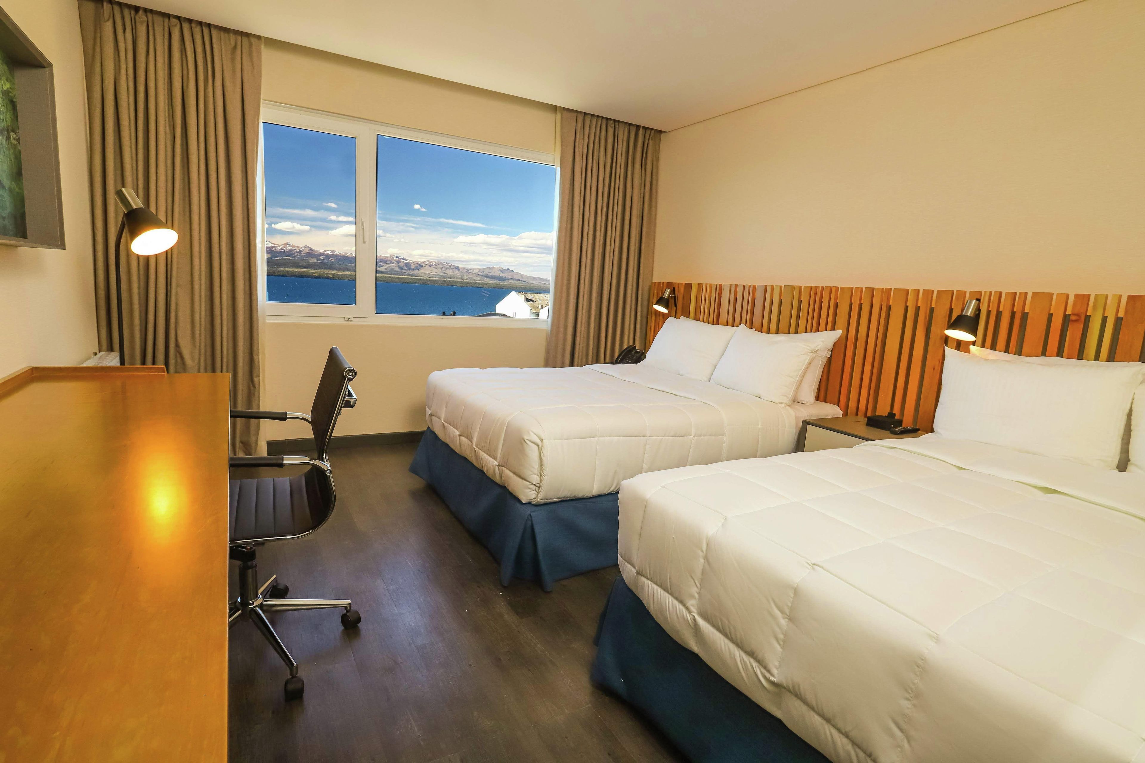 A hotel room with two beds, a desk, a chair, a lamp, and a view of mountains and a lake.