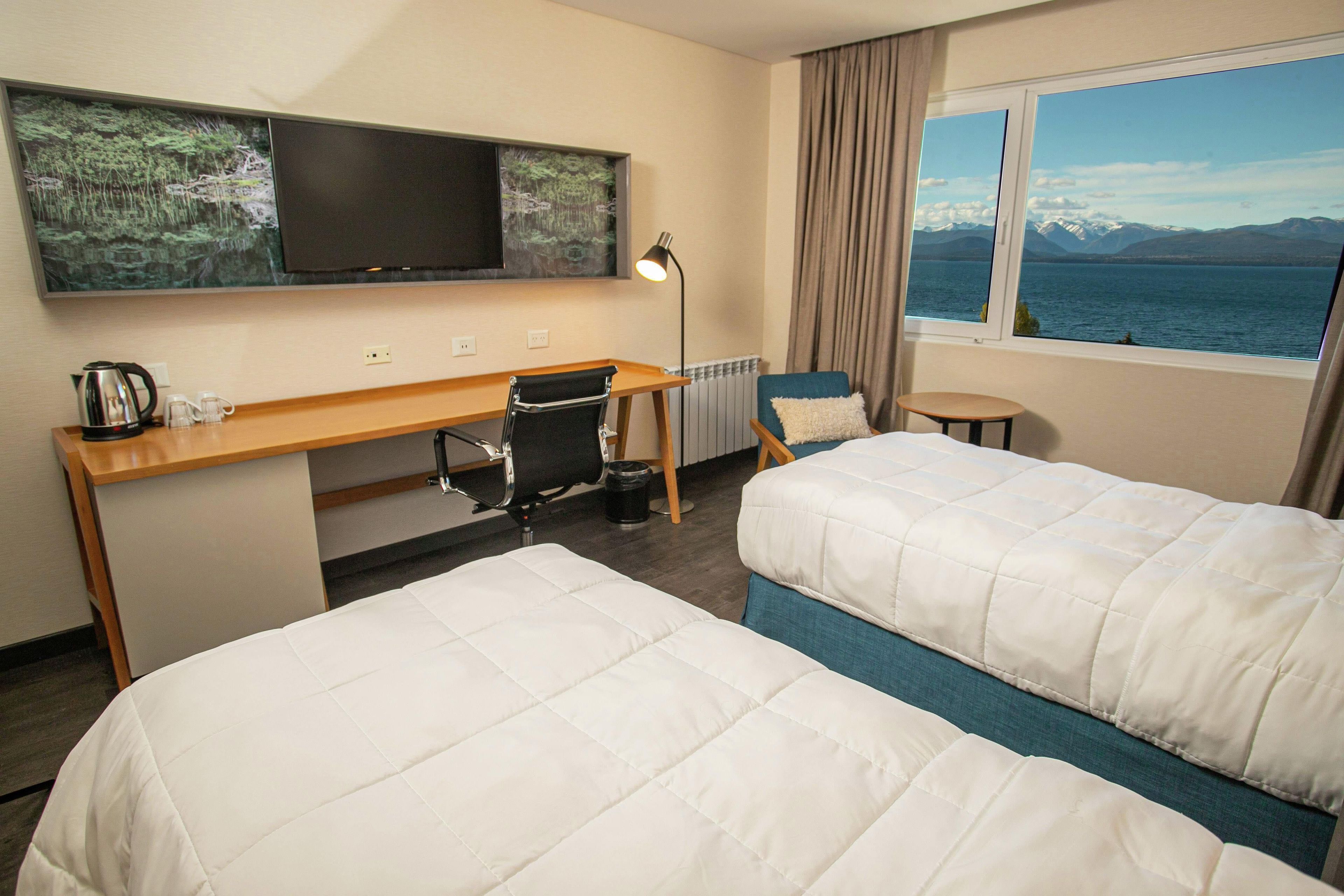 Habitación de hotel con dos camas, un escritorio, una silla, un televisor y una ventana con vista a montañas y al agua.