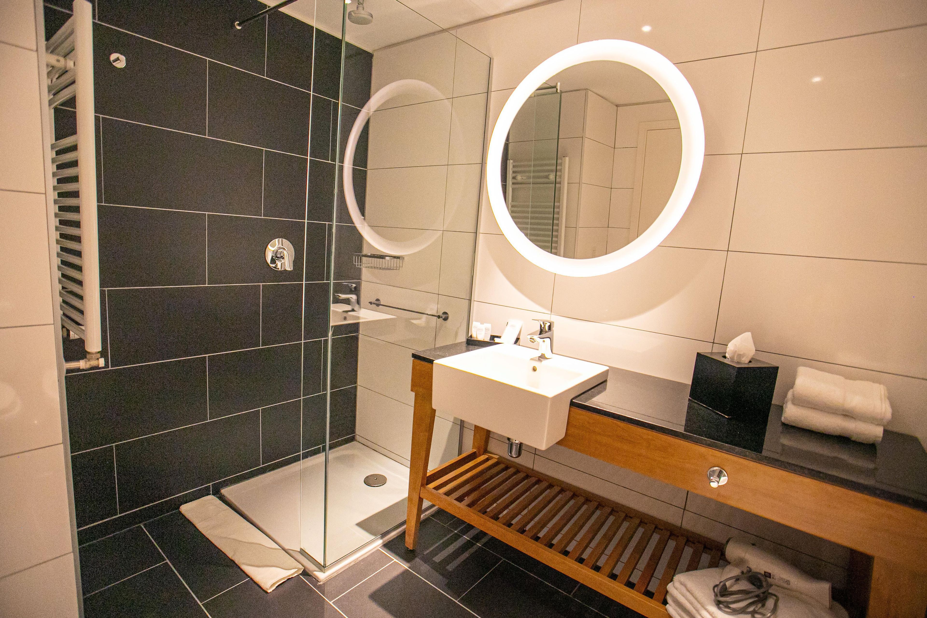 Een moderne badkamer met een douche met glazen wand, een houten wastafelmeubel met een wastafel en twee spiegels aan de muur.