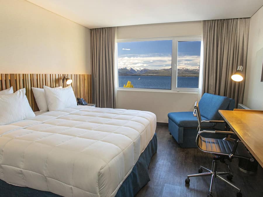 A hotel room with a bed, a desk, a chair, and a window with a view of mountains and a body of water.