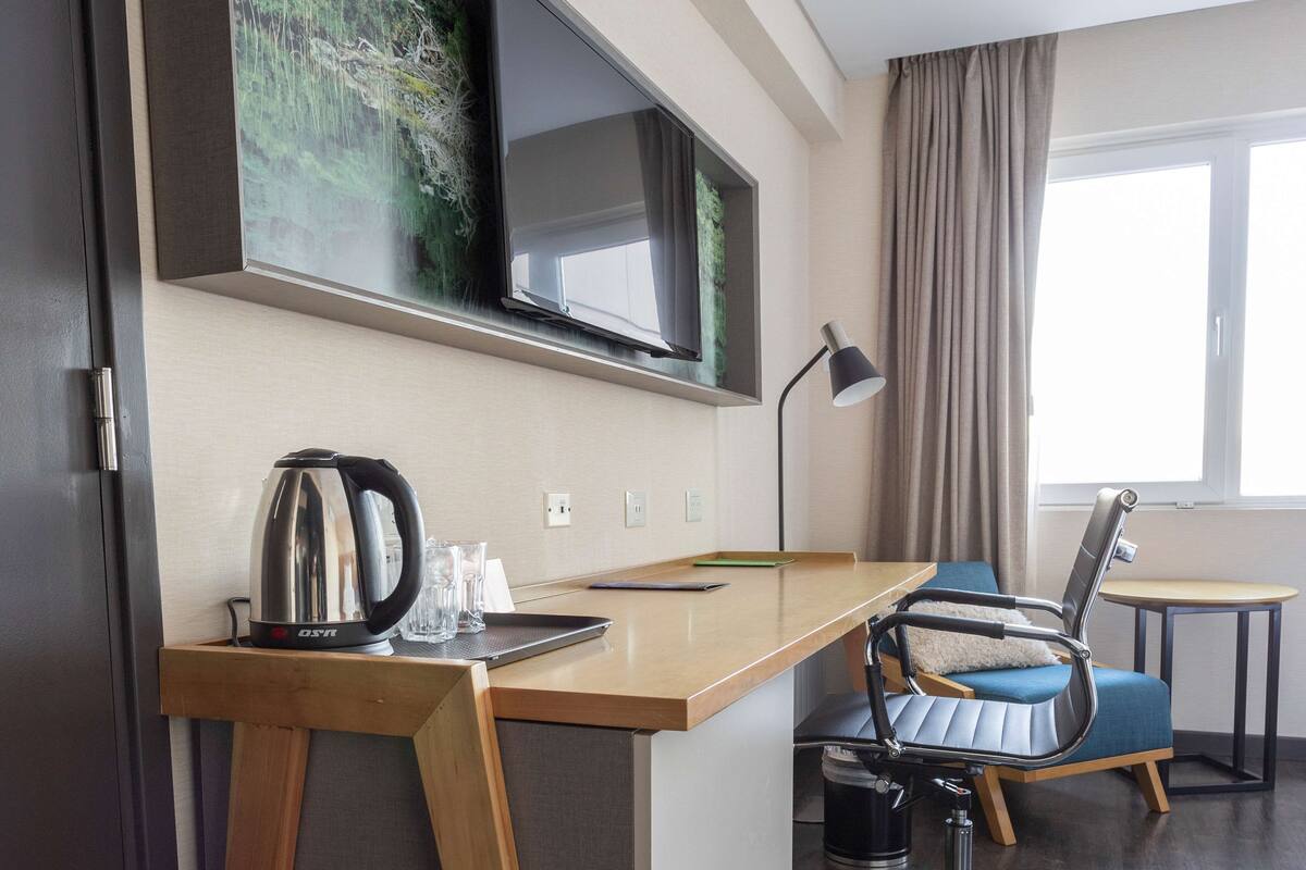 Habitación de hotel con un escritorio de madera, un televisor en la pared, una silla, una mesita y una ventana con cortinas.