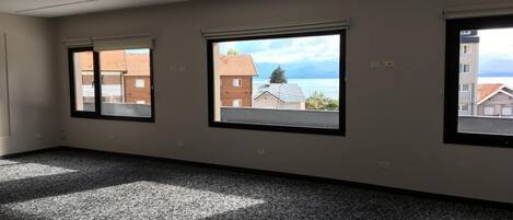 Una habitación amplia con un gran ventanal, piso alfombrado y vistas a edificios y una masa de agua.