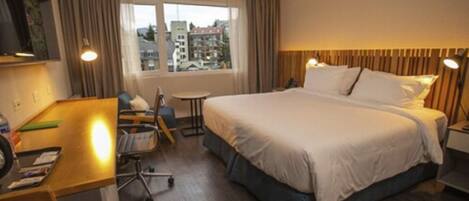 Ein Hotelzimmer mit einem großen Bett, einem Schreibtisch, einem Stuhl und einem kleinen Tisch. Ein Fenster bietet Blick auf Gebäude.