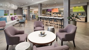 En moderne hotellobby med et siddeområde med runde borde og polstrede stole, et receptionsskranke og et køkkenområde i baggrunden.