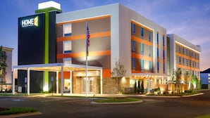 Et moderne hotel med et tydeligt 'HOME 2 Suites by Hilton'-skilt, et flagstang med det amerikanske flag og en godt oplyst indgang.