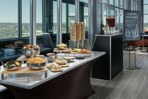 Ein reichhaltiges Buffet mit verschiedenen Speisen und einer Getränkestation in einem modernen Hotelzimmer mit großen Fenstern und Blick auf die Stadt.
