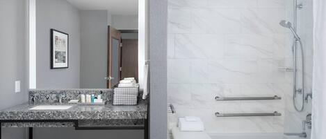 A modern bathroom with a granite countertop, a large mirror, a shower with a glass door, and a small white bench.