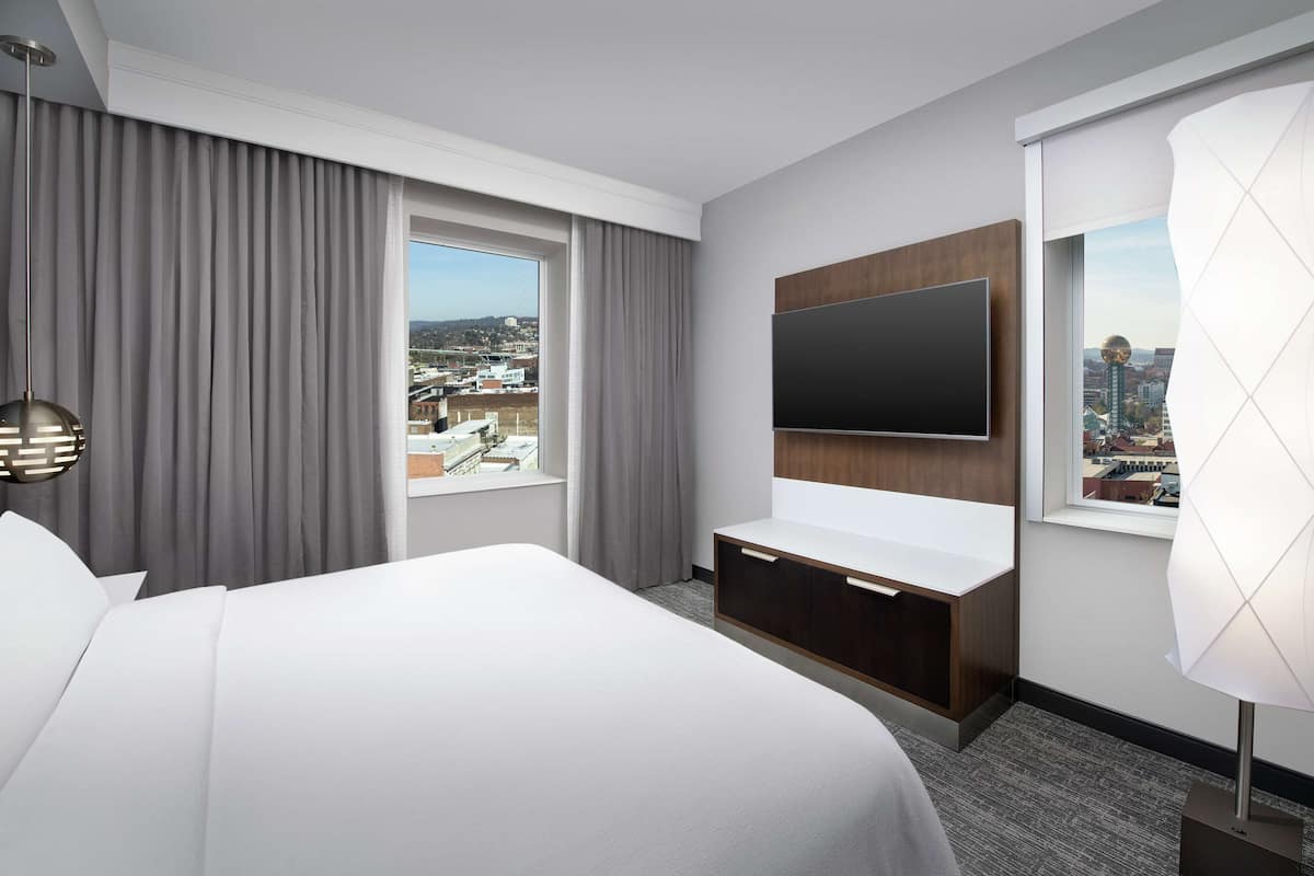 A hotel room with a large bed, a flat-screen TV mounted on the wall, a desk with a chair, and a window with curtains.