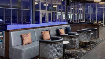 A modern lounge area with a sofa and round tables, large windows with city views, and a blue carpet.