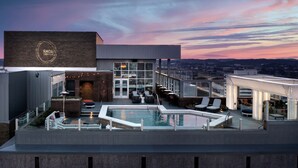 Zona de piscina en la azotea con mobiliario y bar, con vistas a un paisaje urbano al atardecer.