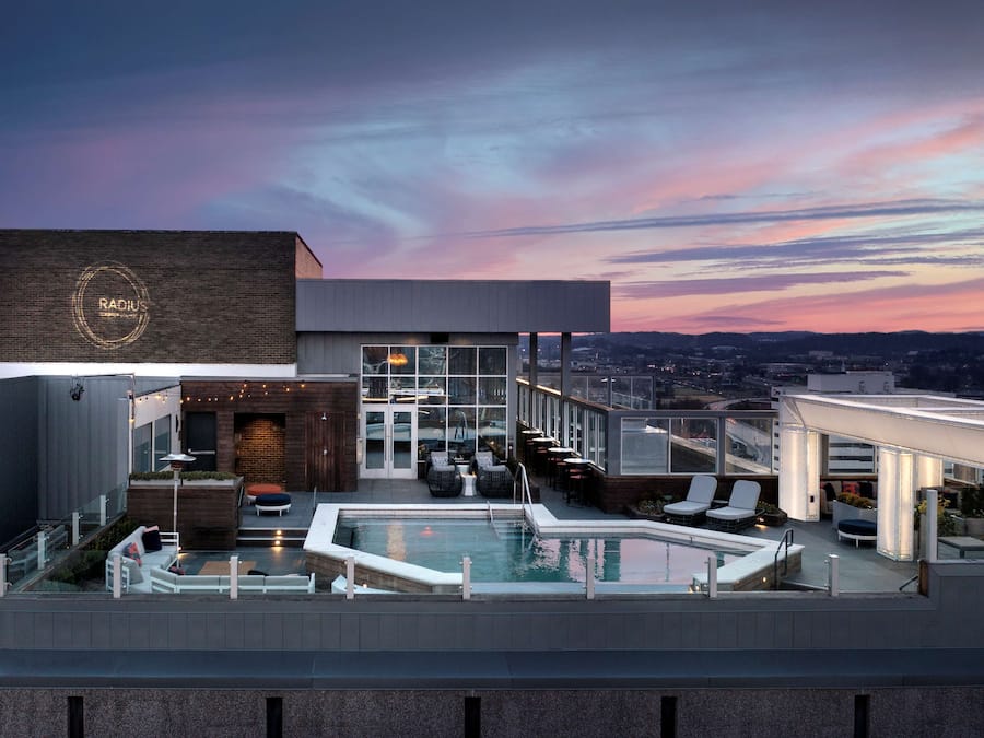 A rooftop pool area with seating and a bar, overlooking a cityscape during sunset.