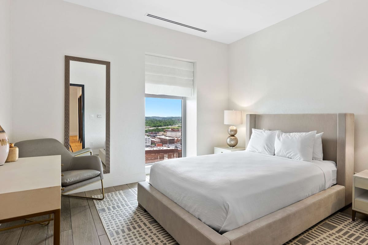 A hotel room with a large bed, two bedside tables with lamps, a desk, a chair, and a large window offering a city view.