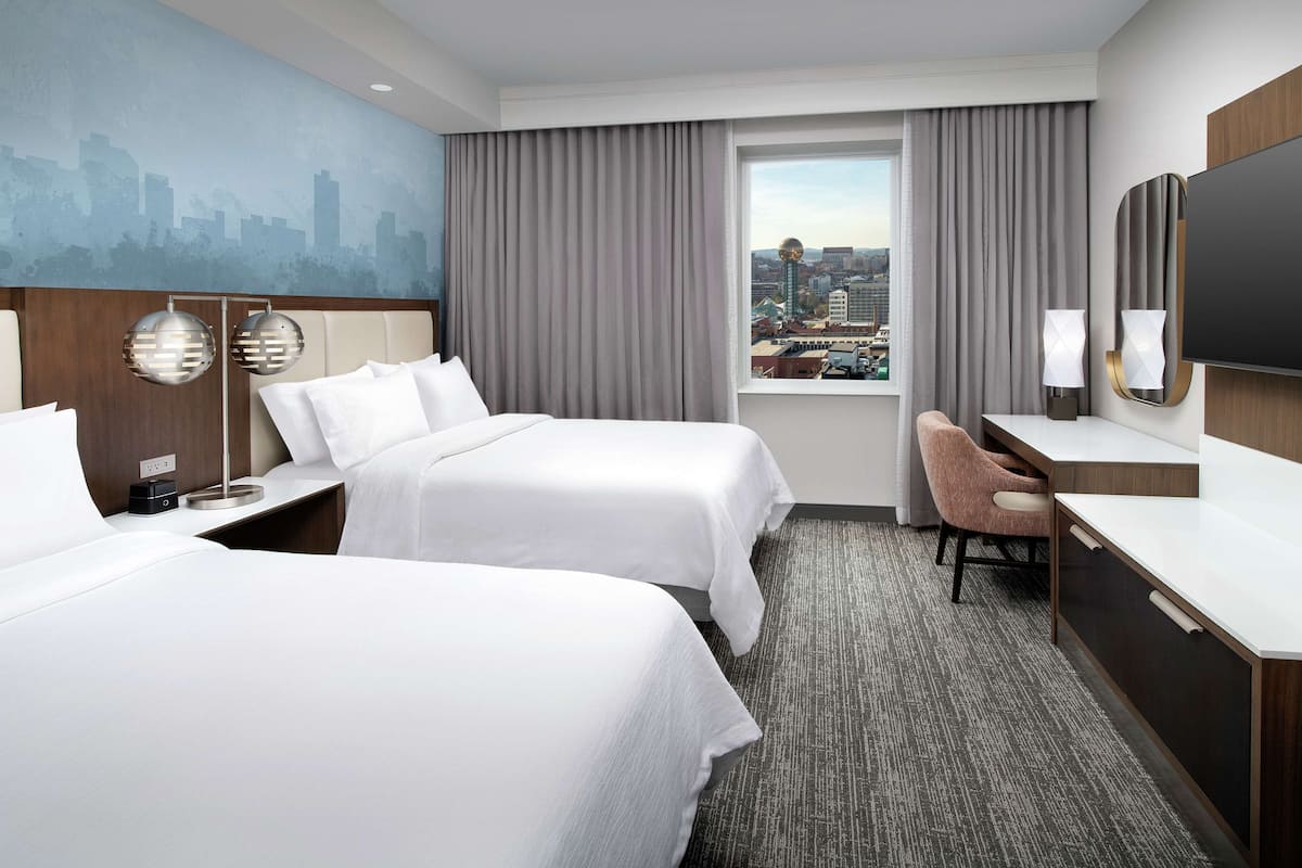 A hotel room with two beds, a desk, a chair, a TV, and a cityscape view.