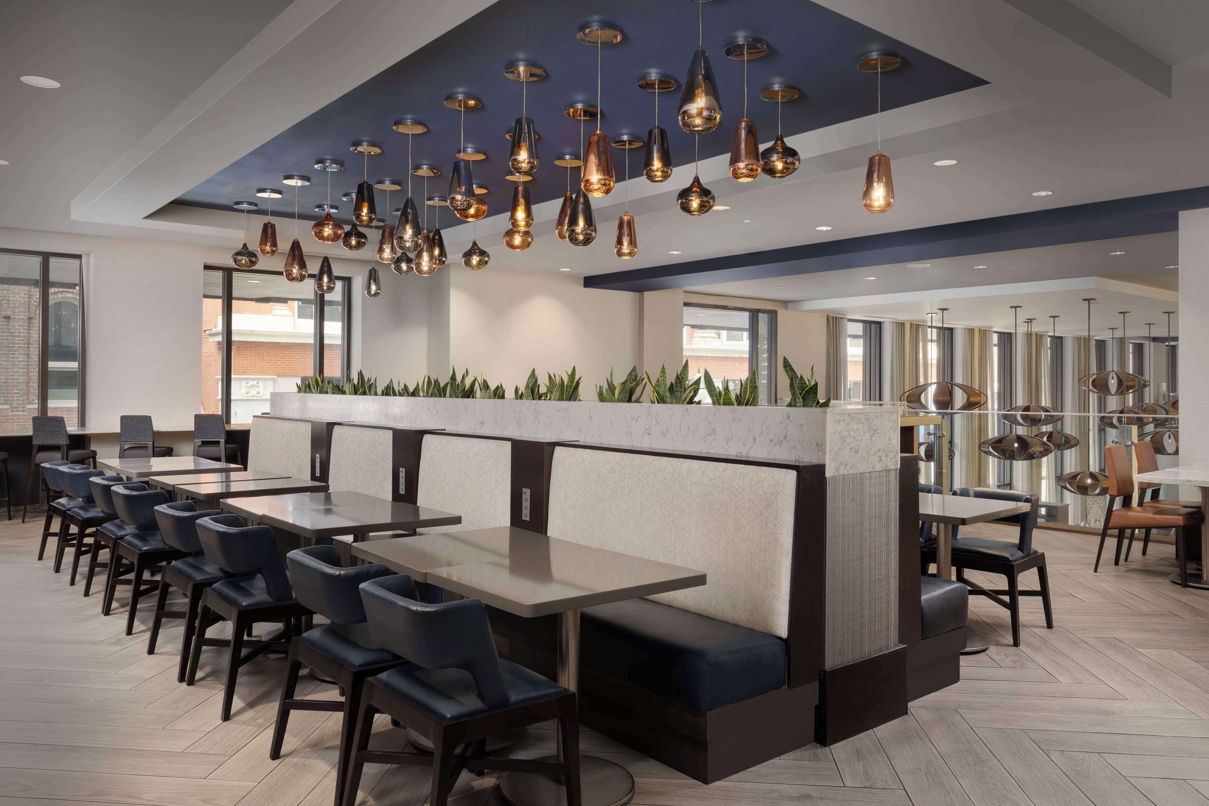 A modern restaurant with multiple tables and booths, pendant lighting, and a view of the cityscape.