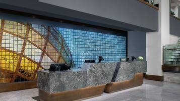A modern hotel reception area with a marble counter, a glass wall with a geometric pattern, and a staircase in the background.