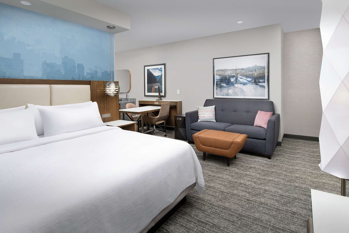 A hotel room with a large bed, a desk, a grey sofa, and a cityscape painting on the wall.