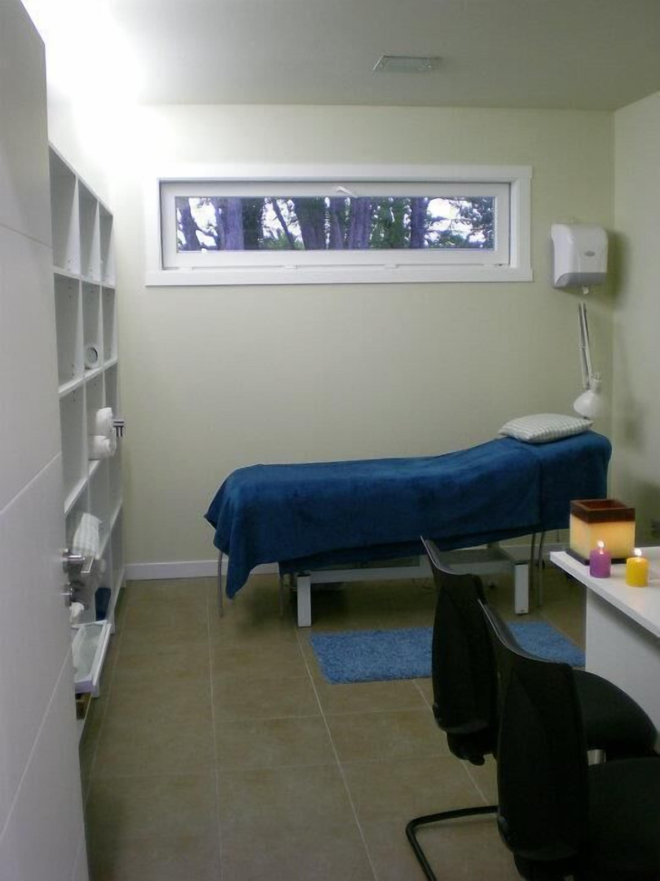 A small, clean room with a single bed, a desk with a chair, a window, and shelves with toiletries.